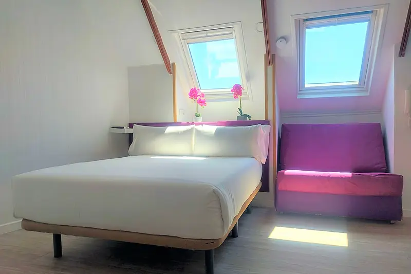 Cama de matrimonio y sofá morado bajo una ventana de techo con luz natural