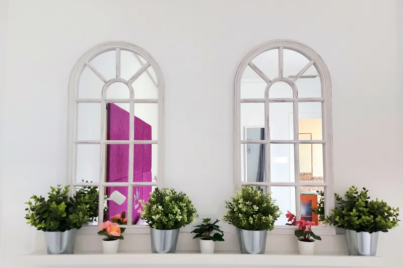 Dos espejos con marco de ventana en arco y plantas sobre una repisa blanca